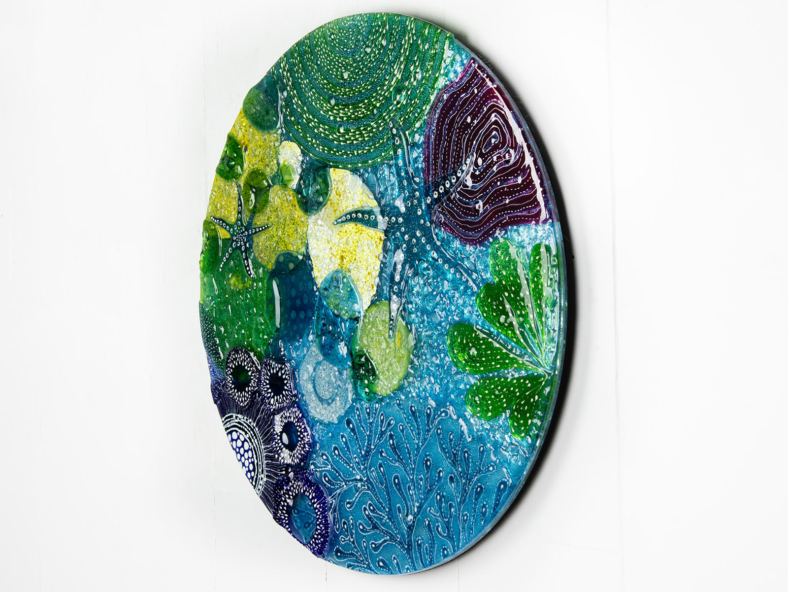 Side on image of round wall panel made from fused glass, inspired by the ocean reefs with starfish detailing and coastal colours. 