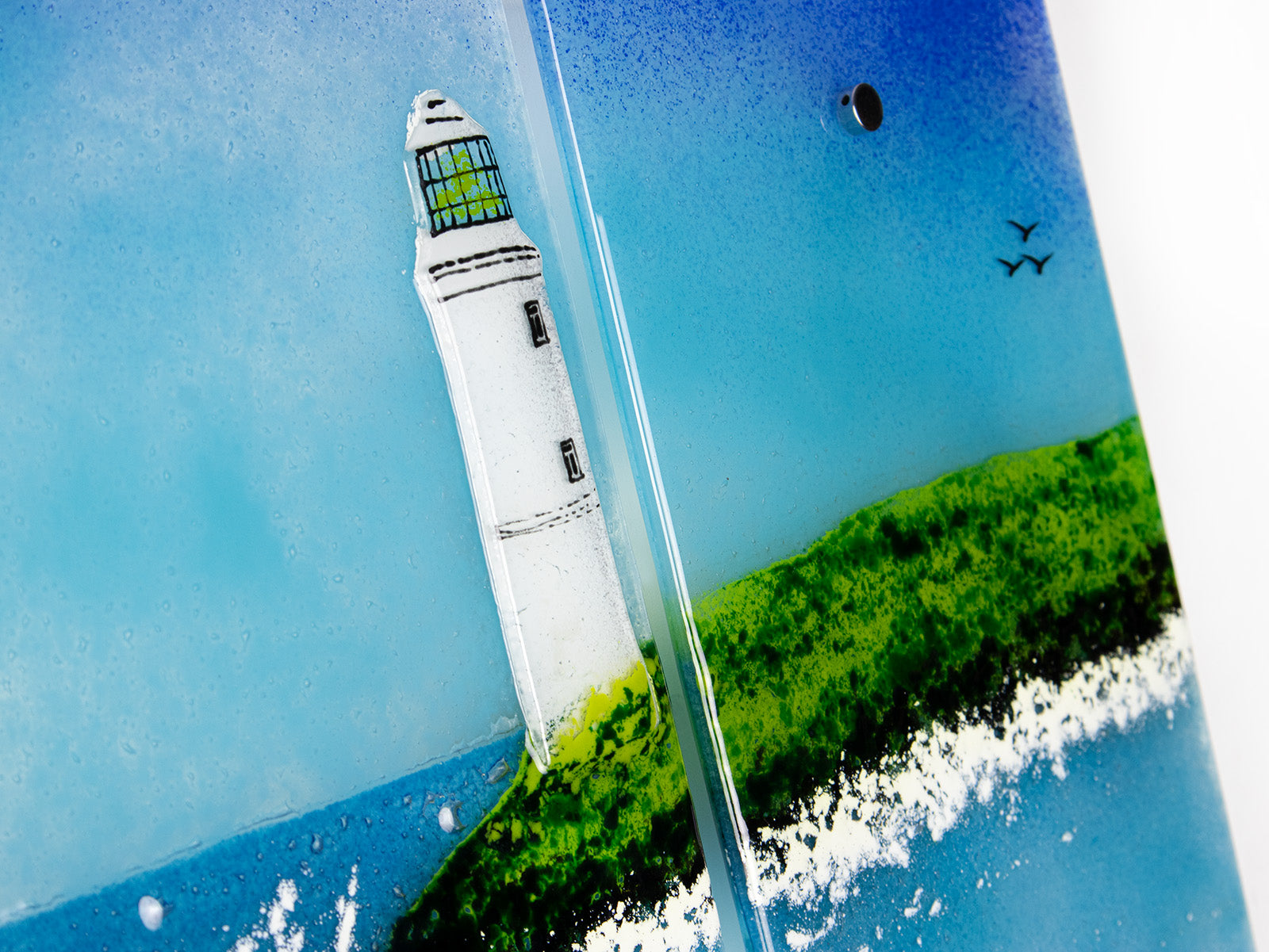 Close up of a lighthouse on a fused glass triptych in coastal colours, with distant soaring sea birds and a Cornish tin mine. Hand-made coastal wall art, with texture. 