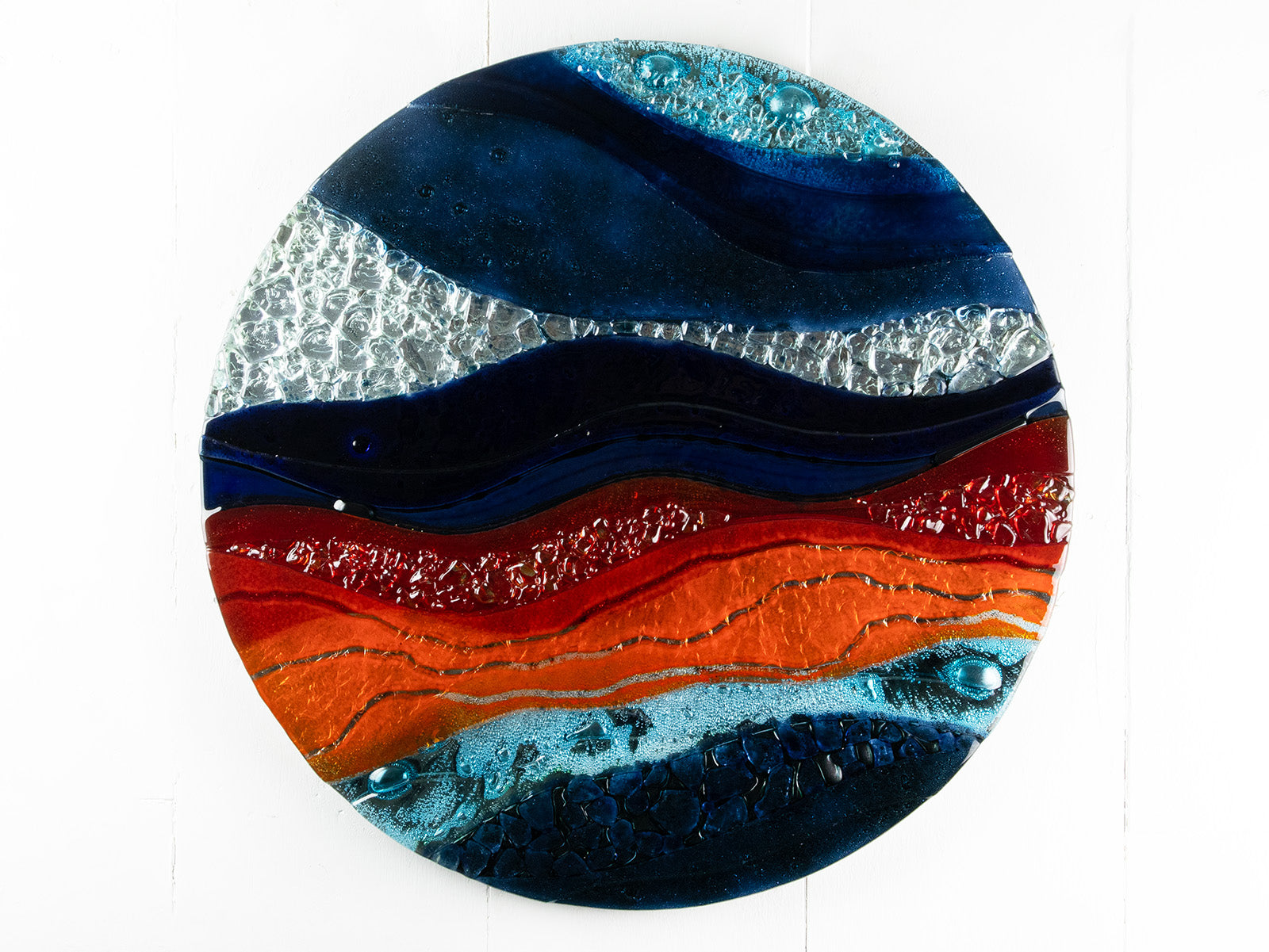 A circular fused glass wall panel featuring abstract wave design in ocean blues and bright oranges, with textured detailing. 