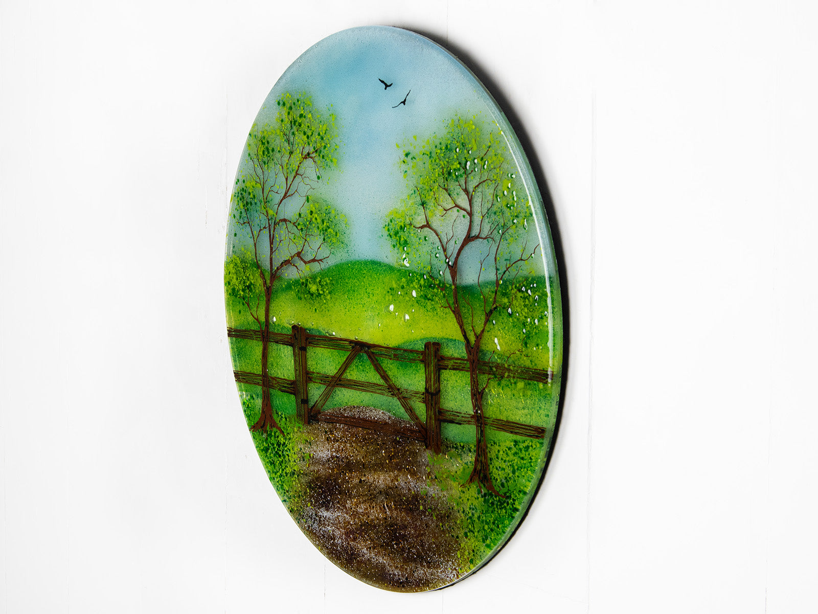 A side view circular fused glass wall art piece featuring a painted landscape with green trees, a wooden fence, rolling hills, and a dirt path under a light blue sky.