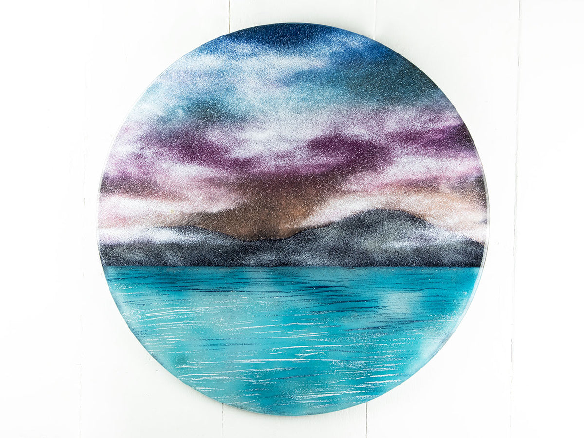 A circular wall art fused glass panel featuring a scenic landscape with a blue and purple sky, misty mountains, and turquoise water. 