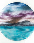 A circular wall art fused glass panel featuring a scenic landscape with a blue and purple sky, misty mountains, and turquoise water. 