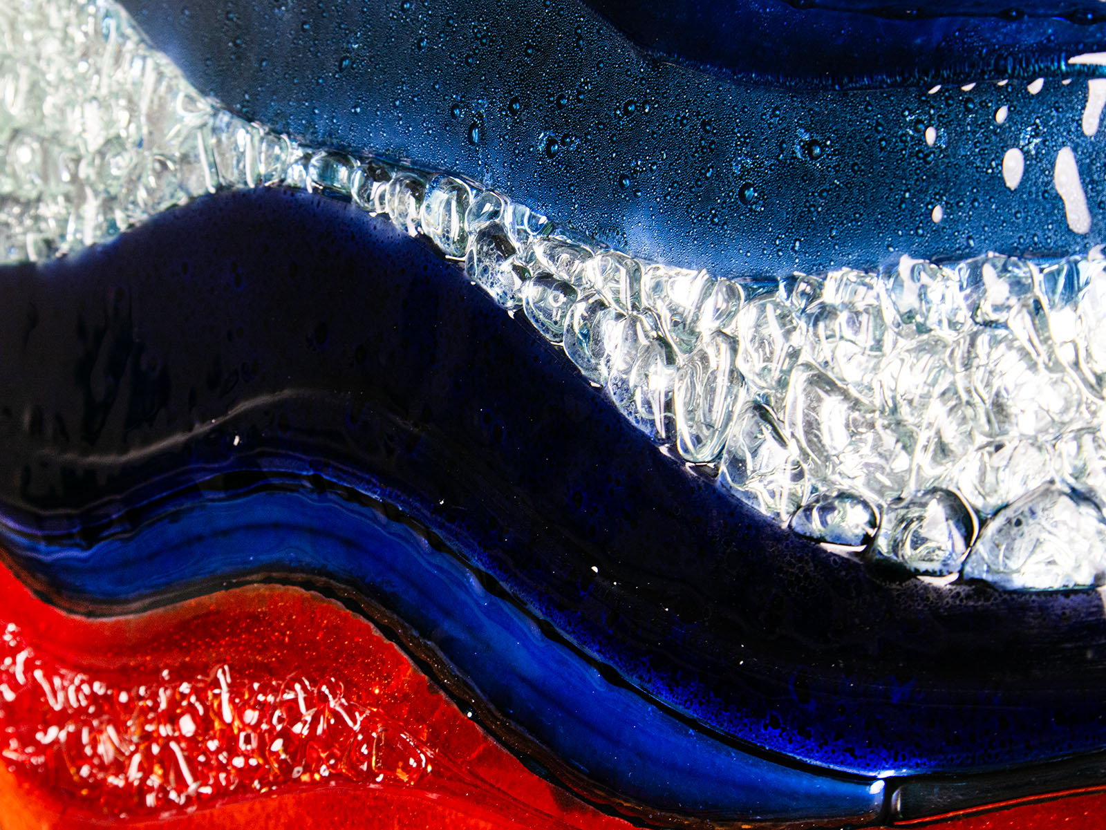 A close up shot of a circular fused glass wall panel featuring abstract wave design in ocean blues and bright oranges, with textured detailing. 