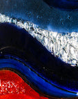 A close up shot of a circular fused glass wall panel featuring abstract wave design in ocean blues and bright oranges, with textured detailing. 