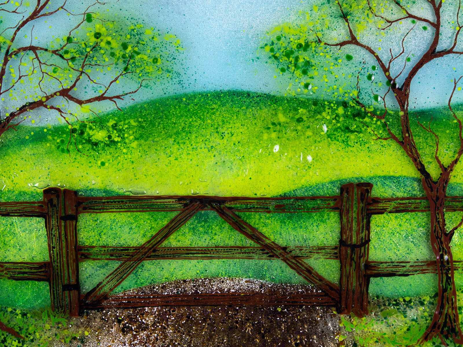 Close up of a circular fused glass wall art piece featuring a painted landscape with green trees, a wooden fence, rolling hills, and a dirt path under a light blue sky.