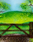 Close up of a circular fused glass wall art piece featuring a painted landscape with green trees, a wooden fence, rolling hills, and a dirt path under a light blue sky.