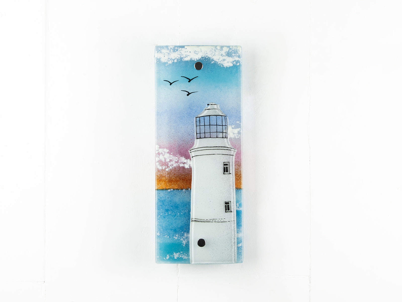 A fused glass extra small wall panel, featuring a lighthouse design with a bright sunset pink sky, with soaring birds. A soft coastal piece of wall art. 