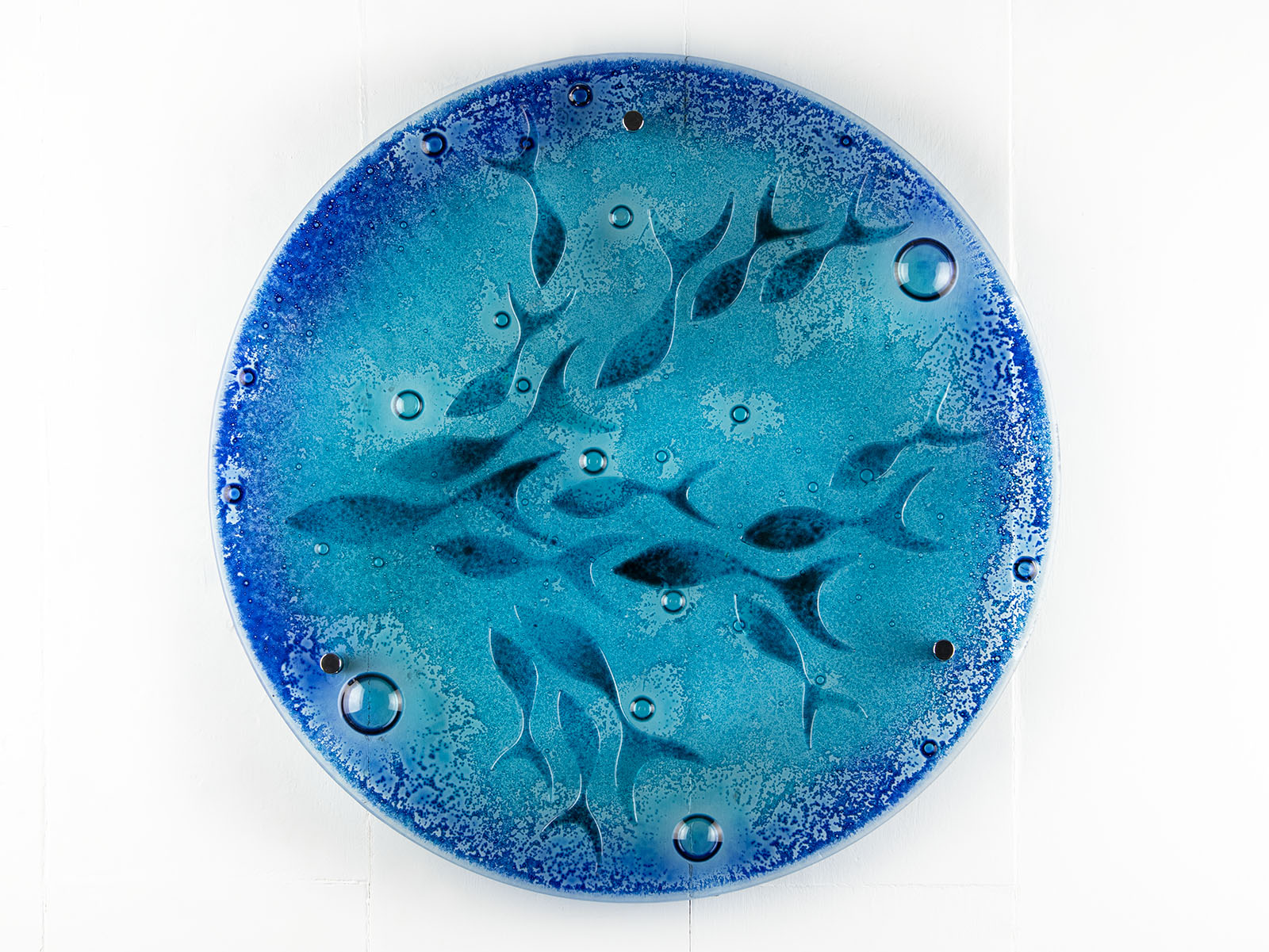 A round fused glass wall panel featuring hand-cut glass fish and bubbles, designed in a marine blue colour-way.