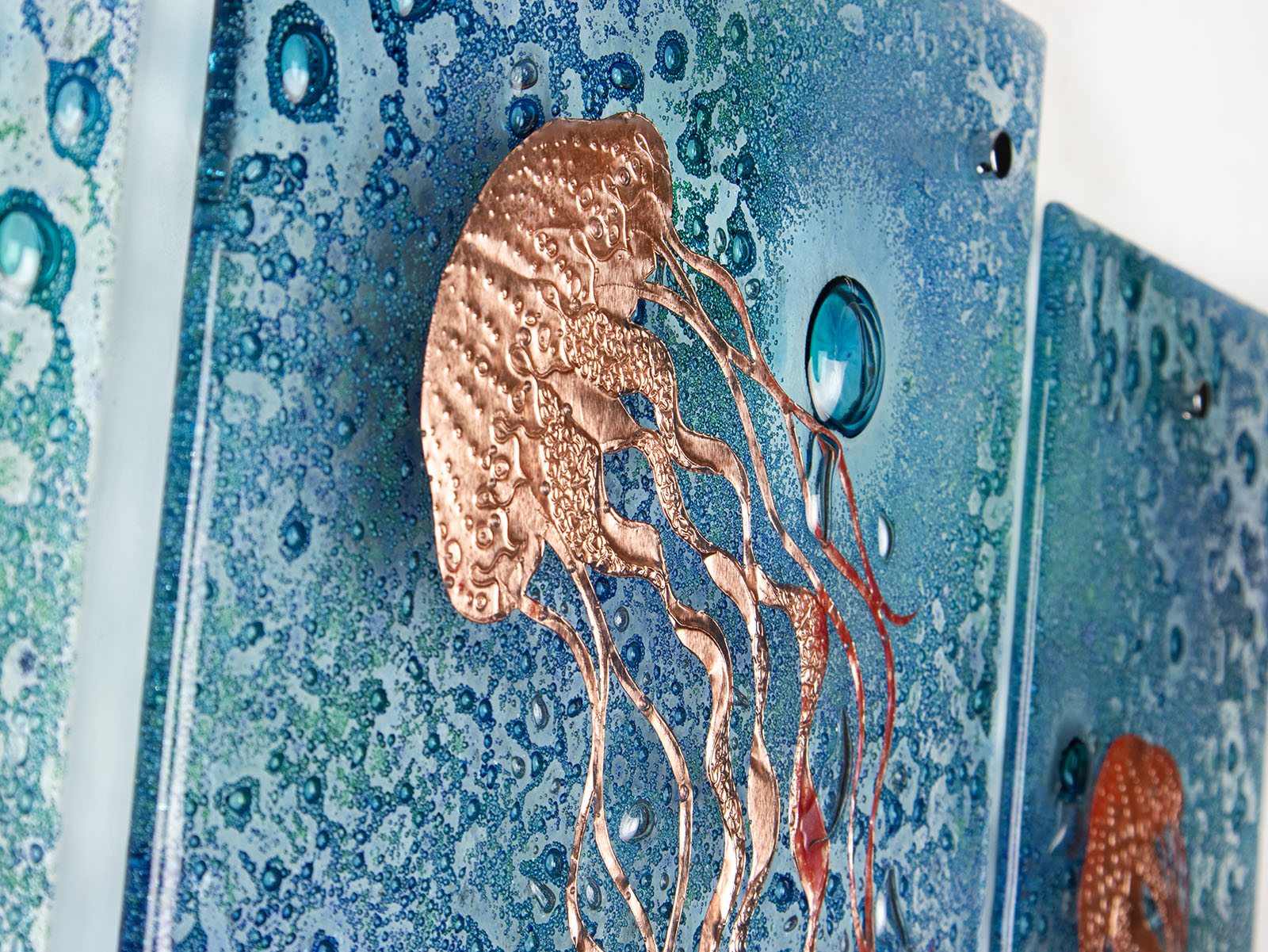 Detailing of fused glass staggered triptych, handmade using copper, which has been hand etched into an intricate Jellyfish design, with bubbled marine blue background. 