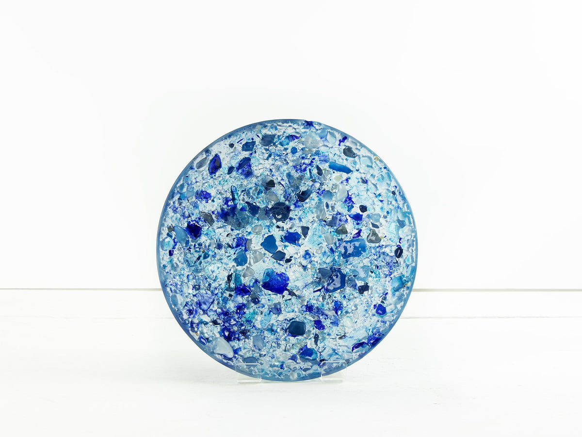 A fused glass round dish, handmade using recycled marine blue hues, designed to create a striking, ocean inspired piece of decor. 