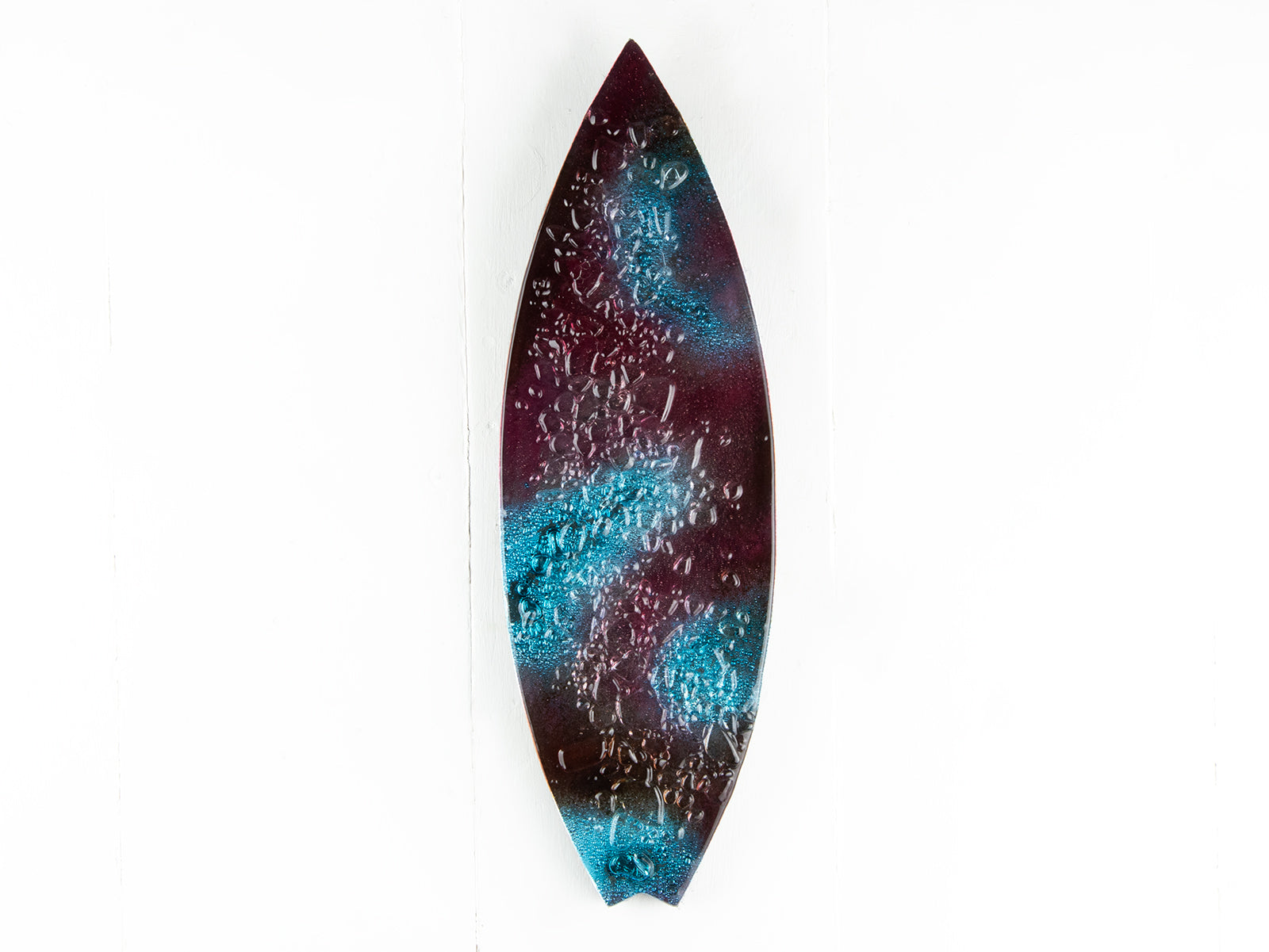 A fused glass surfboard wall panel, handmade in coral pink hues and ocean blues, with textural detailing. A coastal inspired piece of wall art. 