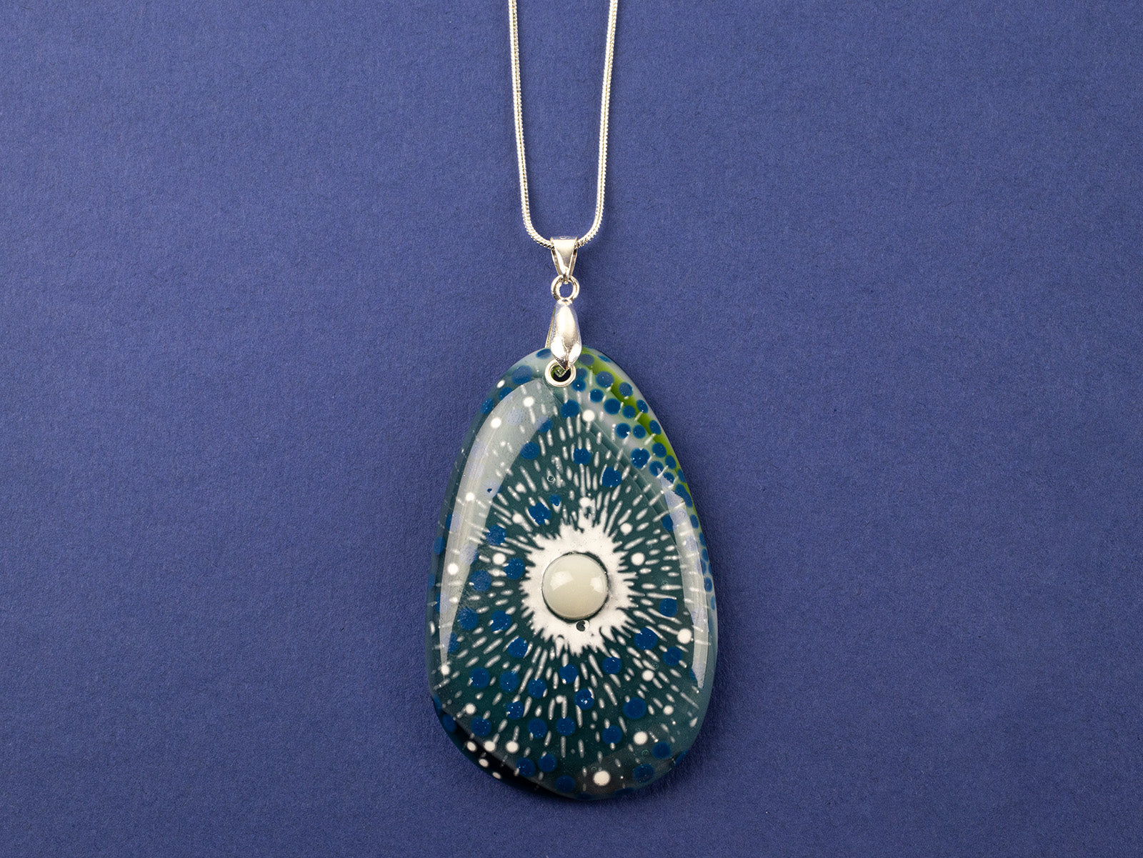 A teardrop-shaped fused glass pendant necklace featuring a blue and green sunburst pattern with white speckles and a central pearl inspired disc, suspended from a sterling silver chain.