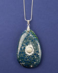 A teardrop-shaped fused glass pendant necklace featuring a blue and green sunburst pattern with white speckles and a central pearl inspired disc, suspended from a sterling silver chain.