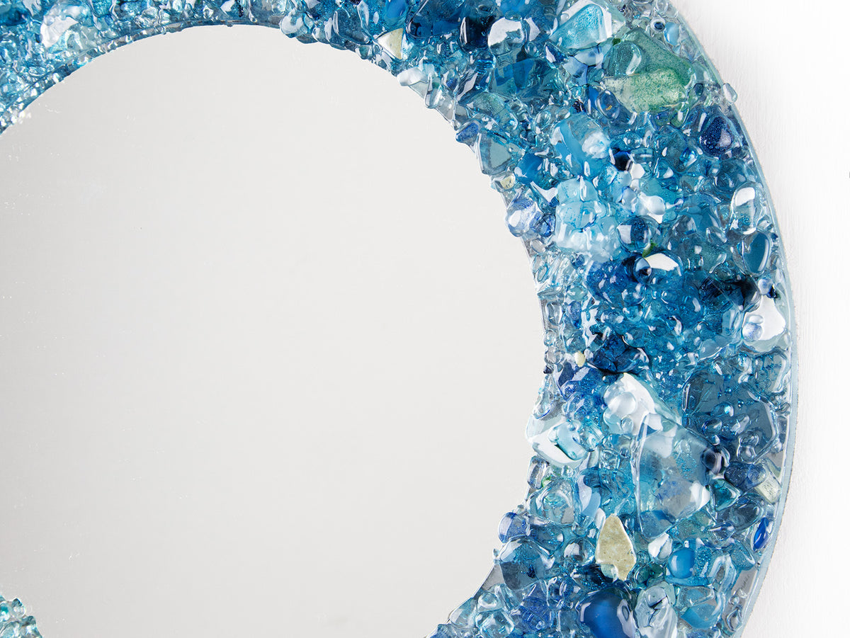 A close up of a fused glass round mirror, handmade using recycled crushed glass in ocean blue hues, with lots of organic texture. 
