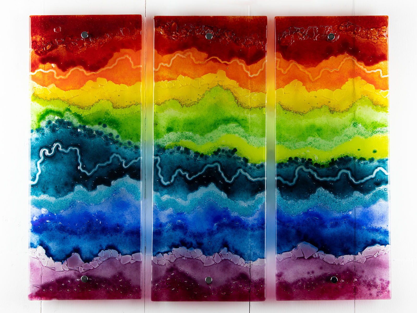 A three-panel vertical fused glass wall art piece featuring wavy horizontal rainbow stripes in red, orange, yellow, green, blue, and purple, mounted on a white wall.