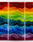 A three-panel vertical fused glass wall art piece featuring wavy horizontal rainbow stripes in red, orange, yellow, green, blue, and purple, mounted on a white wall.