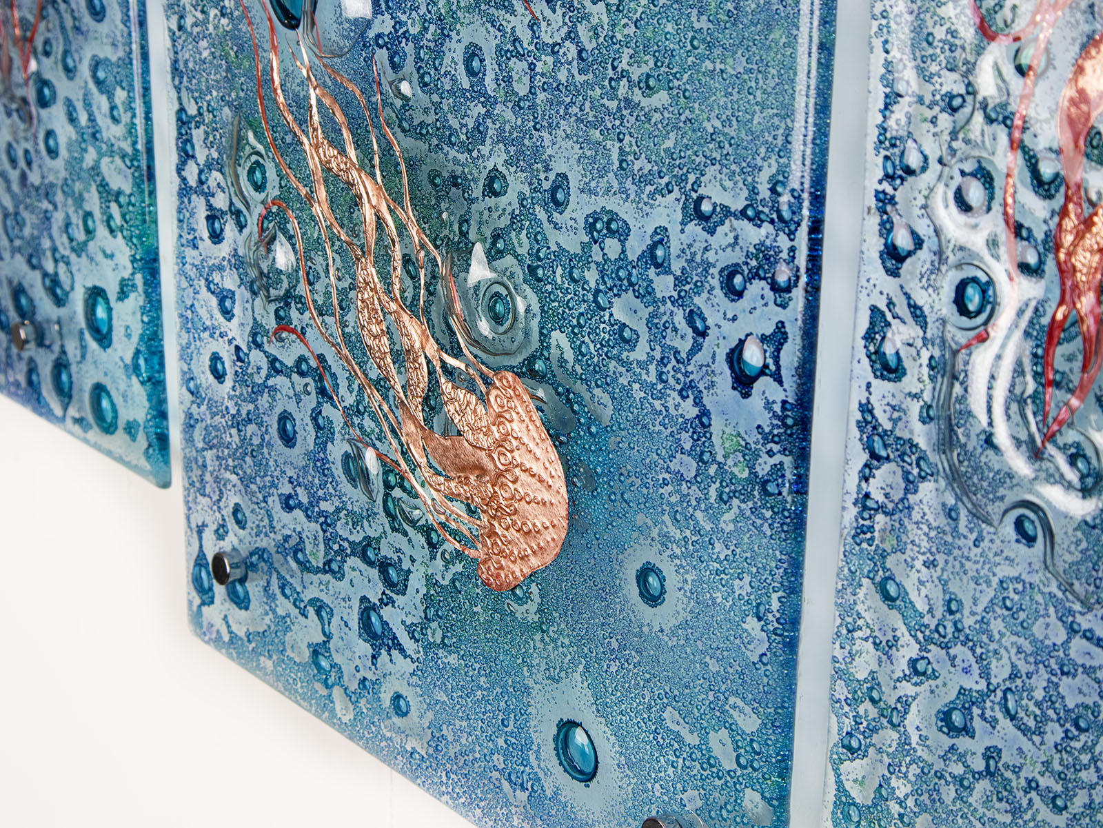 Detailing of fused glass staggered triptych, handmade using copper, which has been hand etched into an intricate Jellyfish design, with bubbled marine blue background. 