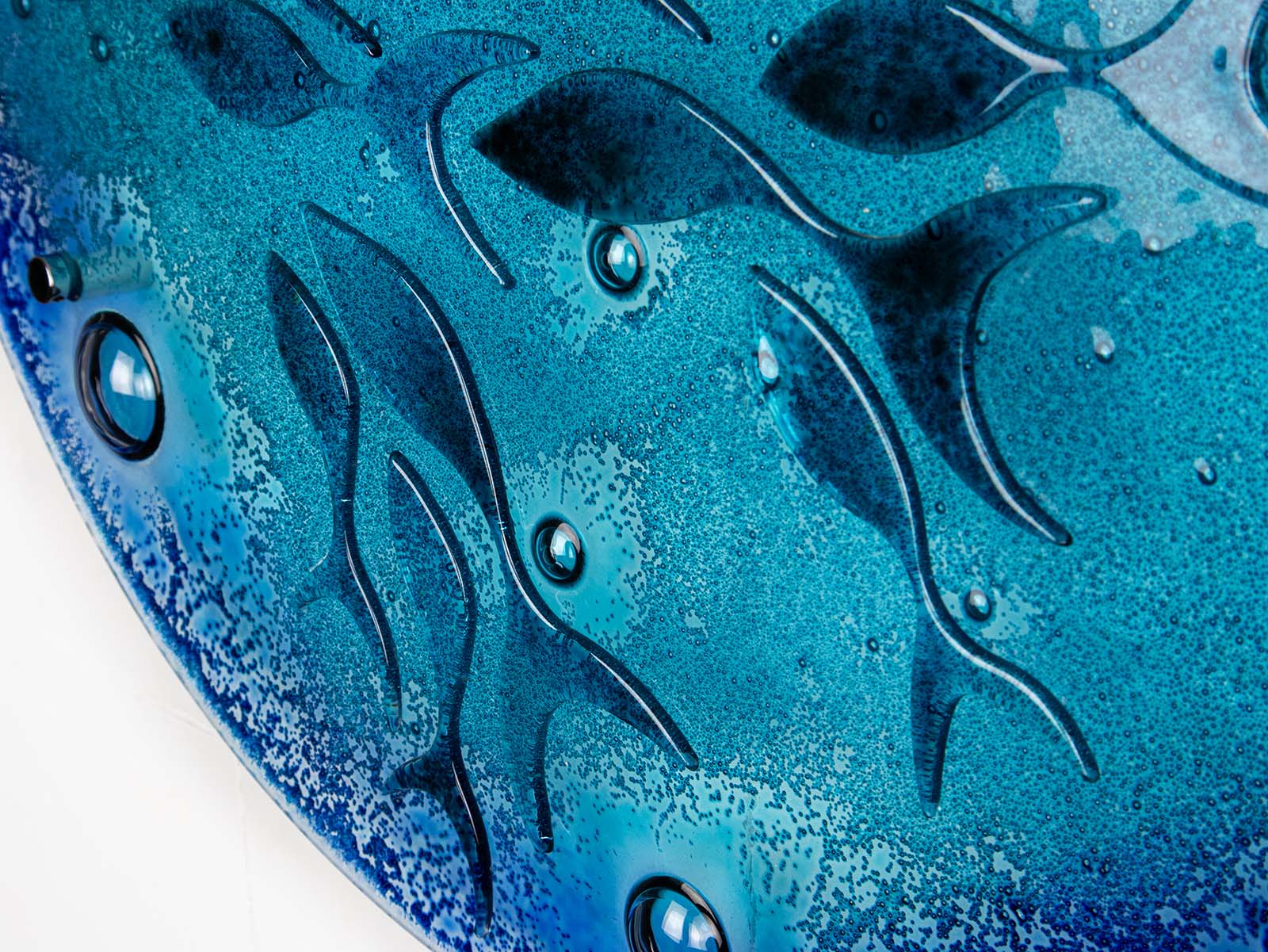A detailed shot of a round fused glass wall panel featuring hand-cut glass fish and bubbles, designed in a marine blue colour-way.