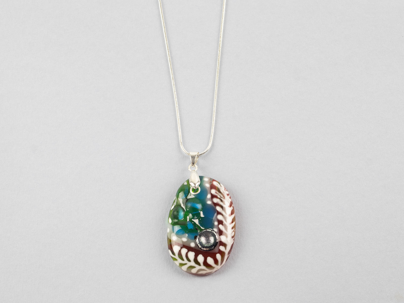 A fused glass pendant necklace on a sterling silver chain, handmade with soft coastal colours and a hint of red, with a hand-painted botanical design and a dichroic glass pebble. 