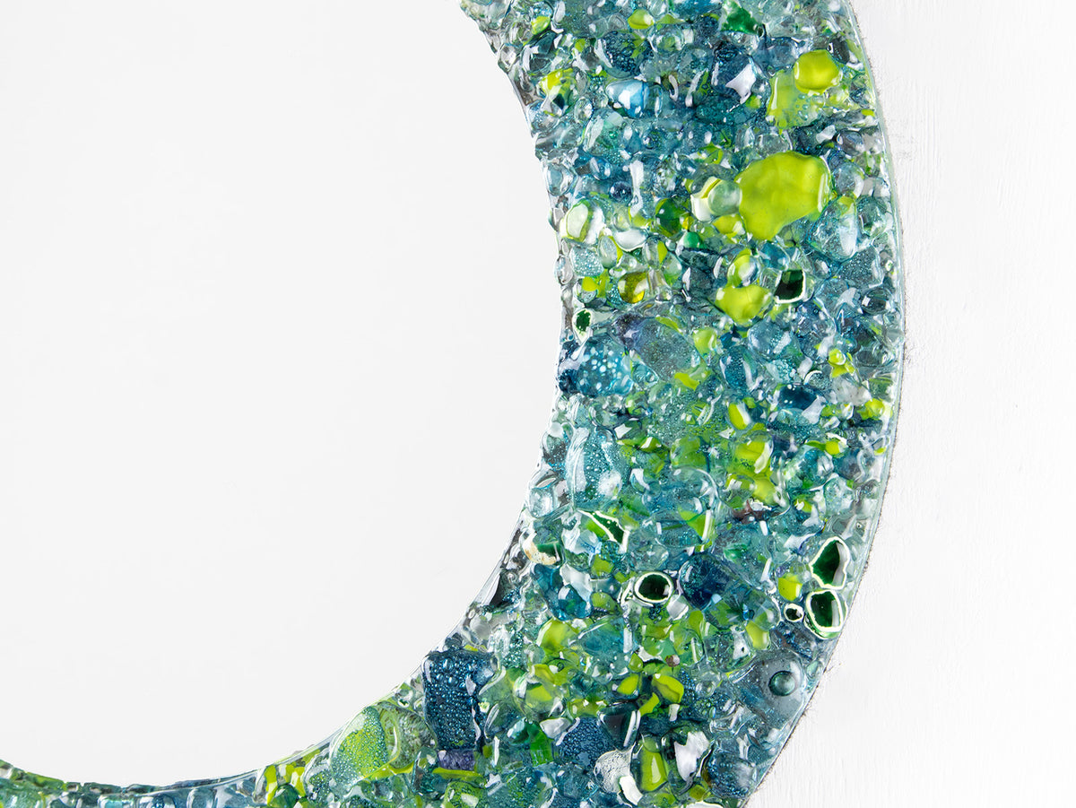 A close up of a fused glass round mirror, handmade using recycled ocean coloured glass in green and aqua hues. A coastal inspired piece of wall art. 