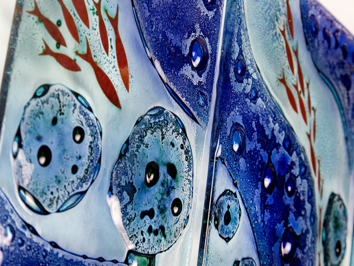 A close up of a fused glass staggered triptych wall display, handmade in bright ocean blues and aquas, in a coastal rock-pool design, featuring copper fish. 