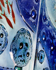 A close up of a fused glass staggered triptych wall display, handmade in bright ocean blues and aquas, in a coastal rock-pool design, featuring copper fish. 