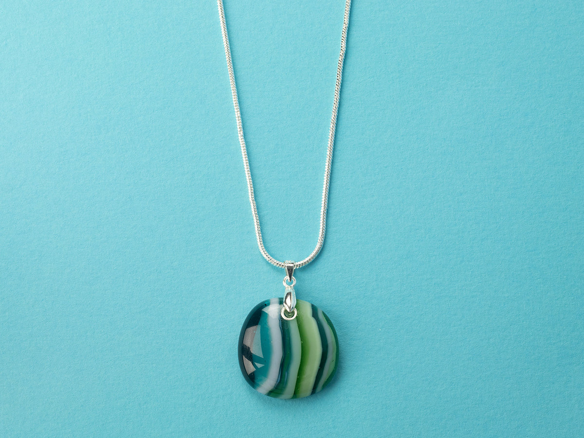 A sterling silver chain necklace with a round fused glass pendant featuring green, teal, and white wavy stripes, handmade in coastal colours. 