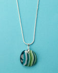A sterling silver chain necklace with a round fused glass pendant featuring green, teal, and white wavy stripes, handmade in coastal colours. 