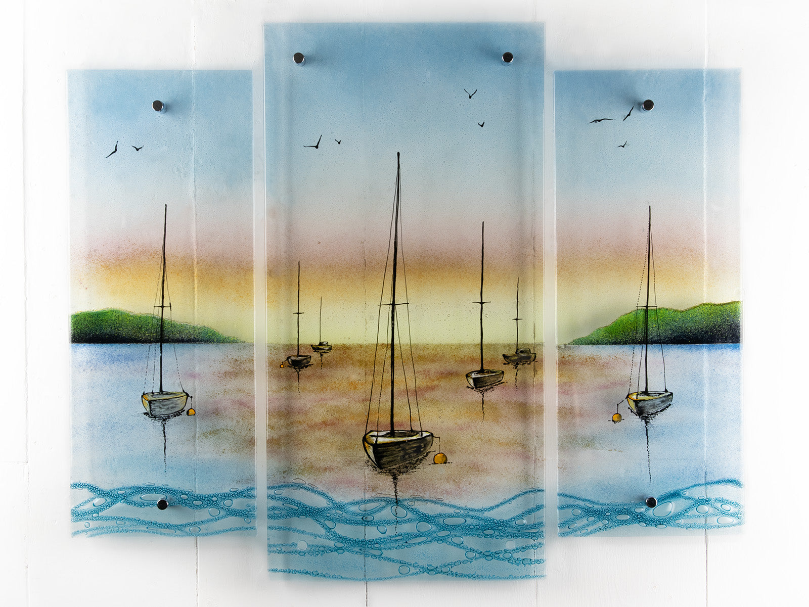 A fused glass triptych depicting a serene harbour scene with hand-painted sailboats on blue water under a pastel sky with hills in the distance.
