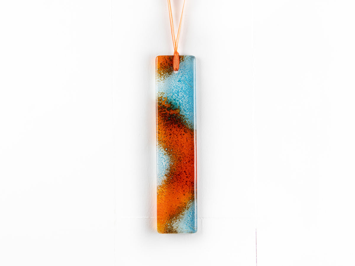 A fused glass hanging, hand-coloured in vibrant orange and bright aqua tones, with an orange ribbon. 
