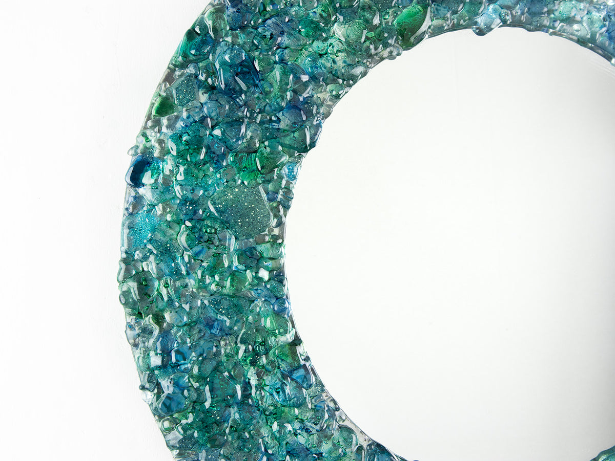A close up of a fused glass round mirror handmade using crushed, recycled glass to create an ocean inspired piece of wall art. 
