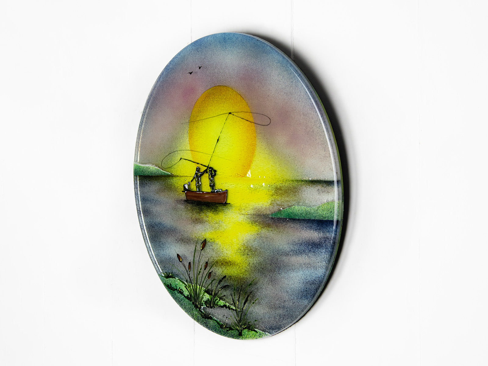A fused glass circular wall art panel featuring a hand-painted design of a fisherman in a small boat casting a line in front of a large glowing yellow sun over a calm lake in natural, organic colours. 
