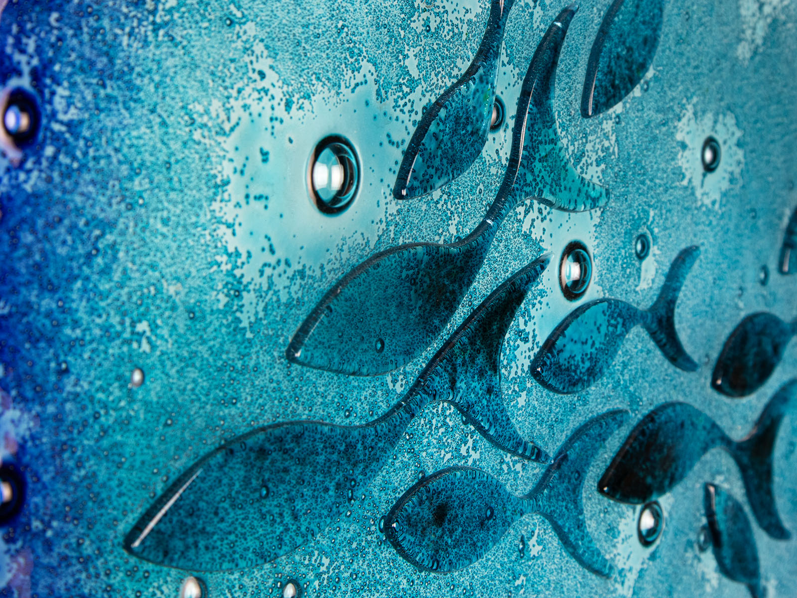 A close up of a round fused glass wall panel featuring hand-cut glass fish and bubbles, designed in a marine blue colour-way.
