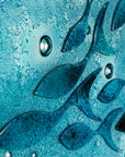 A close up of a round fused glass wall panel featuring hand-cut glass fish and bubbles, designed in a marine blue colour-way.