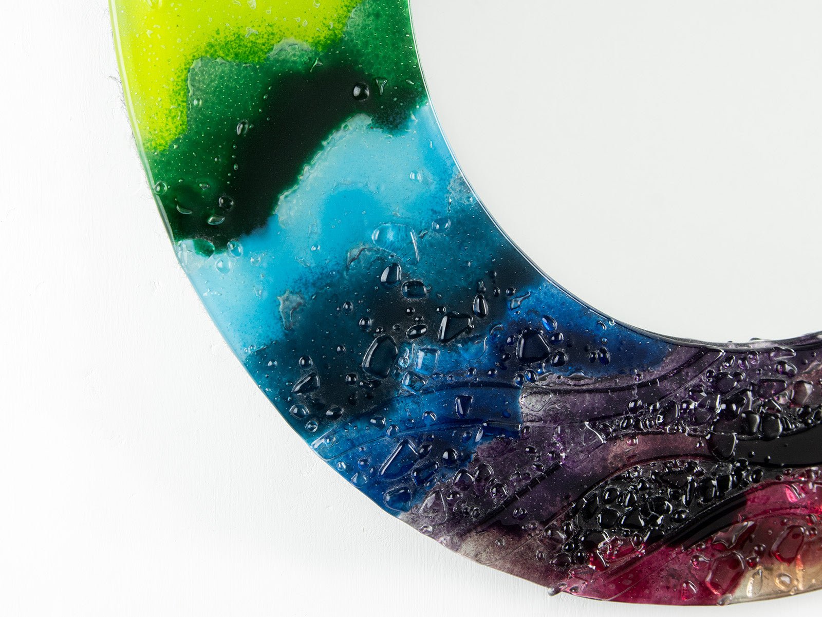 A close up of a circular fused glass round mirror in a vibrant rainbow colour-way with soft waves and textured detailing. 
