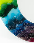 A close up of a circular fused glass round mirror in a vibrant rainbow colour-way with soft waves and textured detailing. 