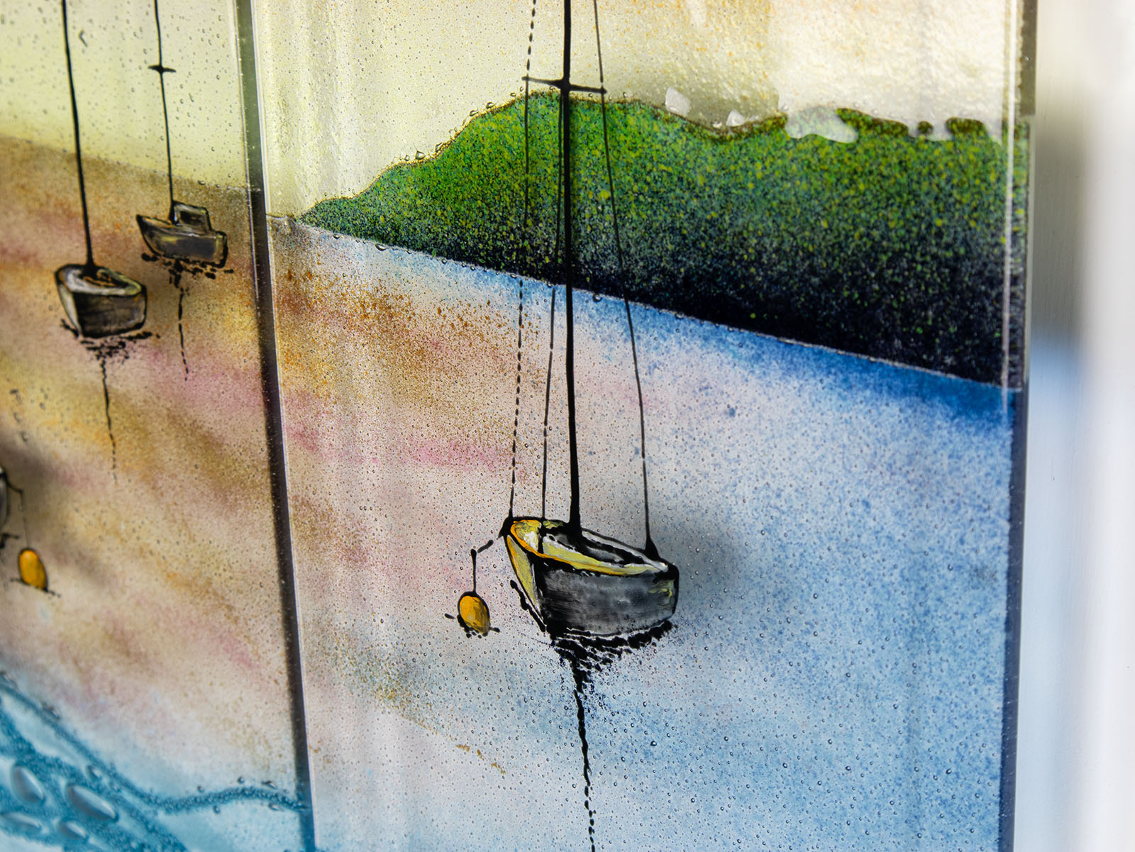 A close up of a fused glass triptych depicting a serene harbour scene with hand-painted sailboats on blue water under a pastel sky with hills in the distance.