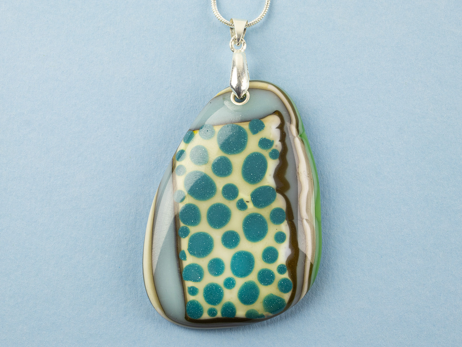 A close up of a handmade fused glass pendant necklace featuring a teardrop-shaped glass charm with a pattern of teal blue dots, bordered by brown and green swirls, suspended from a sterling silver chain.