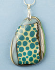A close up of a handmade fused glass pendant necklace featuring a teardrop-shaped glass charm with a pattern of teal blue dots, bordered by brown and green swirls, suspended from a sterling silver chain.