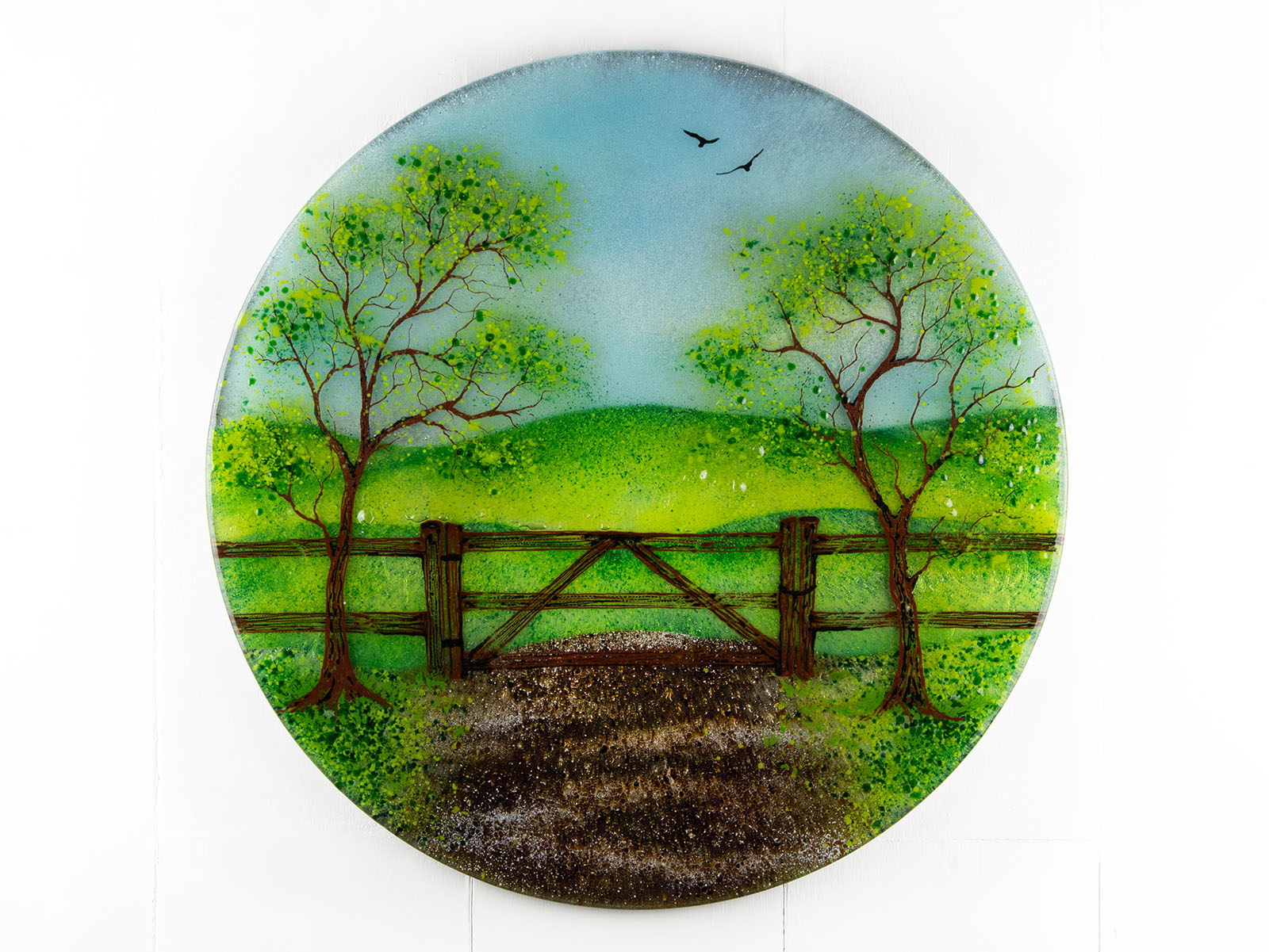 A circular fused glass wall art piece featuring a painted landscape with green trees, a wooden fence, rolling hills, and a dirt path under a light blue sky.