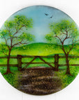 A circular fused glass wall art piece featuring a painted landscape with green trees, a wooden fence, rolling hills, and a dirt path under a light blue sky.