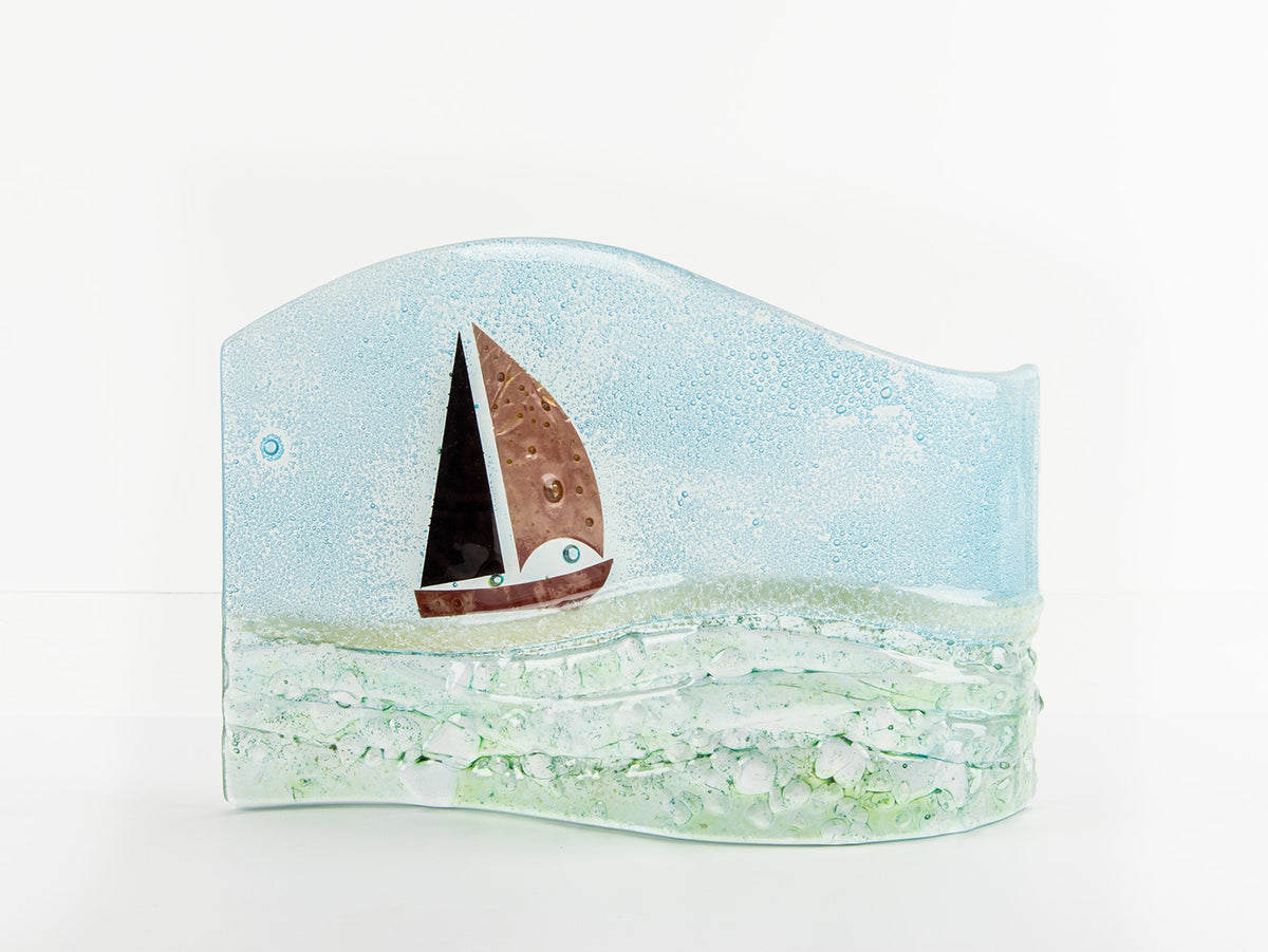 A hand fused glass wave sculpture with a metal sail boat and a soft coastal colour-way, with textured details. 