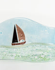 A hand fused glass wave sculpture with a metal sail boat and a soft coastal colour-way, with textured details. 