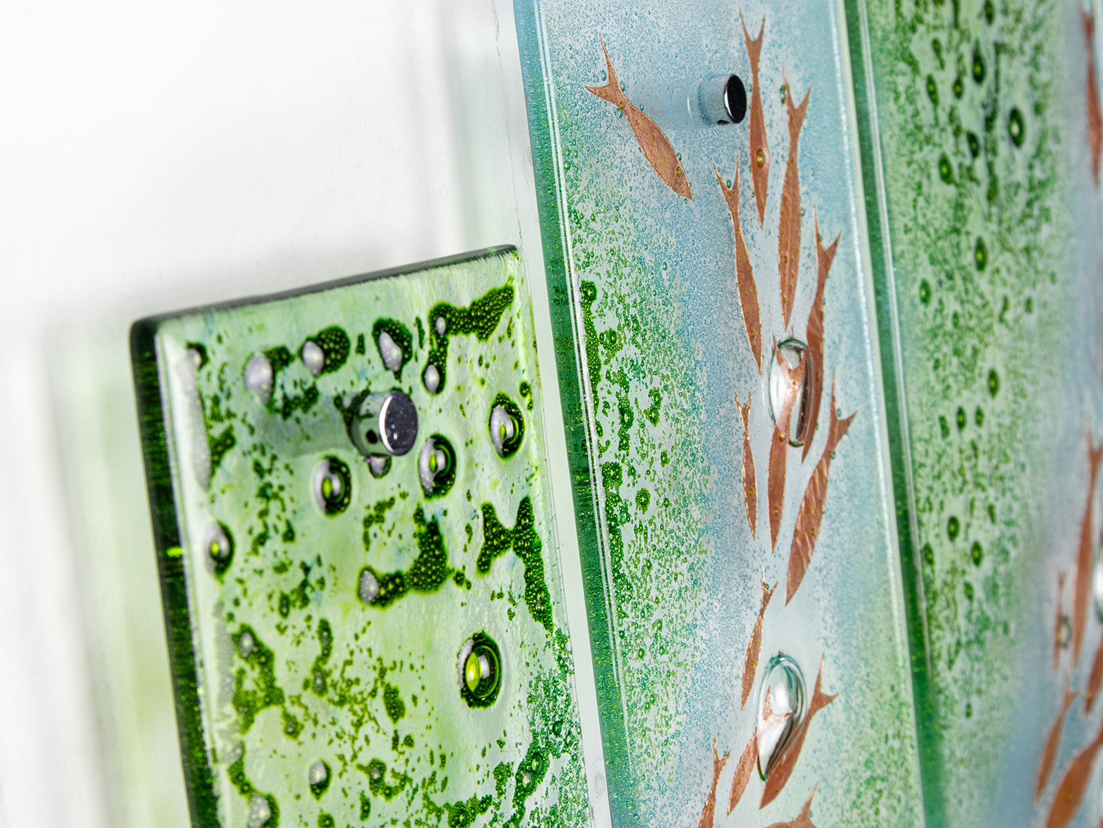 A detailed shot of a fused glass quintych featuring a soft coastal colour-way with darting copper fish and bubbled detailing. An ocean inspired piece of wall art. 
