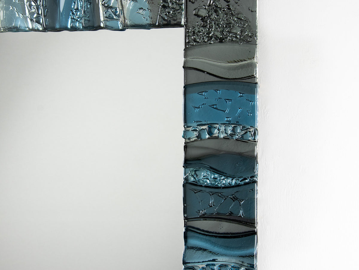 A close up of a fused glass oblong mirror, handmade using soft coastal blue tinted and grey glass with delicate texture and a wavy design.